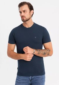 Granatowa koszulka z krótkim rękawem wykonana z miękkiego materiału, z okrągłym dekoltem i małym logo na piersi. Noszona z dżinsami.