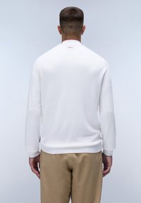 Uomo visto da dietro che indossa una camicia bianca a maniche lunghe e pantaloni beige, su uno sfondo chiaro e uniforme.