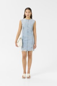Vestido de ganga, azul claro, sem mangas, ajustado com bainha desfiada e costuras visíveis. Acessoriado com uma mala de cor creme e sandálias de salto.