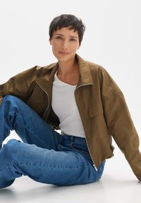 Braune leichte Jacke mit Reißverschluss, zwei Brusttaschen und entspanntem Schnitt, kombiniert mit einem weißen T-Shirt und blauen hoch taillierten Jeans.