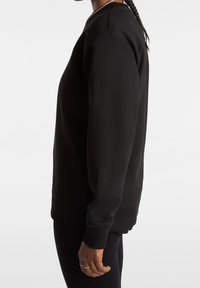 Person i svart långärmad sweatshirt och svarta byxor med flätat hår, sedd från sidan mot en helt vit bakgrund.