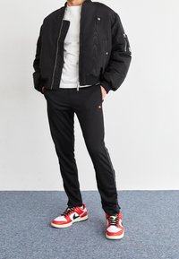 Zwarte bomberjack met ritssluiting, witte T-shirt eronder, zwarte broek met zijkantdetails en rood-witte sneakers met zwarte accenten.