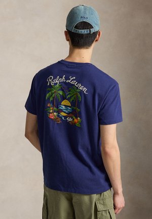 Mann trägt ein blaues Ralph Lauren T-Shirt mit tropischem Palmen- und Sonnenuntergangs-Stick, grüne Cargoshorts und eine hellblaue Polo-Kappe, gesehen von hinten.