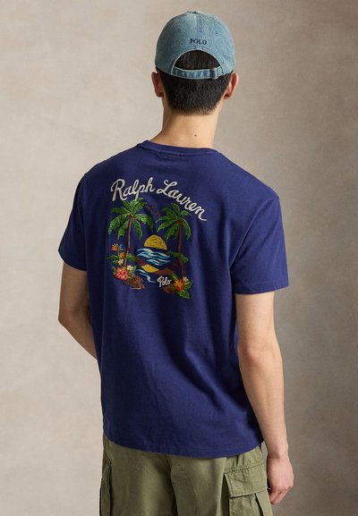 Άνδρας φοράει μπλε μπλουζάκι Ralph Lauren με κέντημα τροπικής φοινικιάς και ηλιοβασιλέματος, πράσινα cargo σορτς και ανοιχτό μπλε καπέλο Polo, κοιτάζοντας από πίσω.