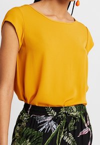Žlutý krátký rukáv top vyrobený z lehké tkaniny s kulatým výstřihem, v kombinaci s černými kalhotami s tropickým zeleným a růžovým vzorem.