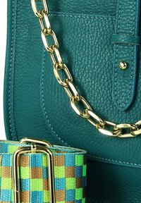 Tegelkleurige leren handtas met een gestructureerde afwerking, een gouden kettingbandje en een gevlochten band met groene, blauwe en bruine geruite patronen.