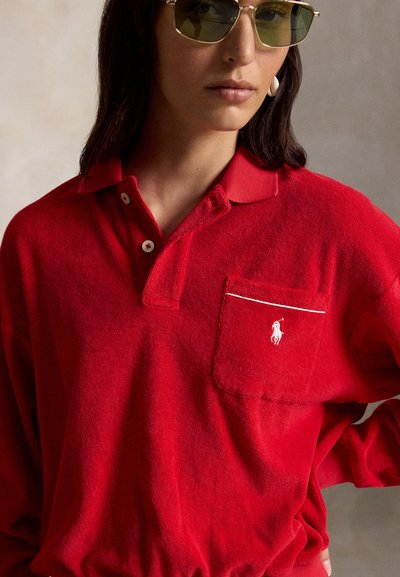 Femme portant des lunettes de soleil à verres teintés verts et un polo rouge à manches longues avec un logo blanc brodé sur la poche poitrine.