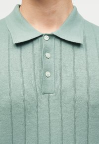 Κοντινή λήψη ανοιχτοπράσινου πλεκτού πόλο με υφή, τρία λευκά κουμπιά και διπλωμένο γιακά στο πάνω μέρος του στήθους και του λαιμού ενός ατόμου.