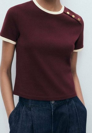 Mujer lleva una camiseta de manga corta burdeos con ribete crema y tres botones dorados en el hombro, combinada con pantalones de cintura alta azul oscuro.