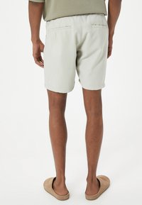 Hombre con pantalones cortos beige claros y zapatos slip-on marrones de pie con la espalda hacia la cámara sobre un fondo blanco.