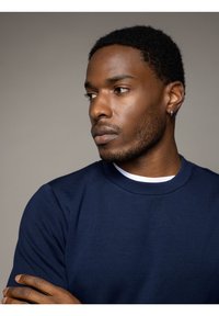 Maglietta maniche corte blu navy con scollatura rotonda, indossata sopra una camicia bianca. Il tessuto sembra morbido e il modello ha un orecchino.