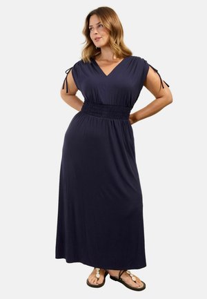 Moteris dėvi be rankovių tamsiai mėlyną maxi suknelę su prigludusia juosmens linija ir raišteliais ant pečių, derina su juodomis sandalėmis su baltais karoliukais.