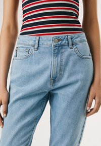 Jeans de mezclilla de color azul claro con una cintura alta, pierna recta, bolsillos frontales y cierre de botón metálico. Combinados con una blusa sin mangas a rayas.