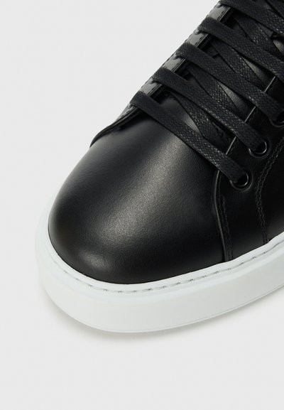Baskets en cuir noir avec une finition lisse, bout rond et lacets noirs texturés. Semelle en caoutchouc blanc avec des détails de couture le long du bord.