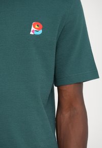 Tumši teal krāsu īsu piedurkņu polo krekls ar teksturētu audumu. Tam ir daudzkrāsains izšūts logotips uz krūšu daļas. Taisna apakša.