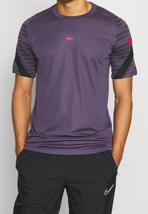 Mand iført lilla atletisk t-shirt med korte ærmer og mønstrede ærmer samt sorte atletikbukser med hvidt logo, hænderne i lommerne.