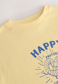 Maglietta gialla in cotone con una grafica blu che raffigura un pretzel e il testo "HAPPY PRETZEL." Girocollo e maniche corte. Tessuto liscio.