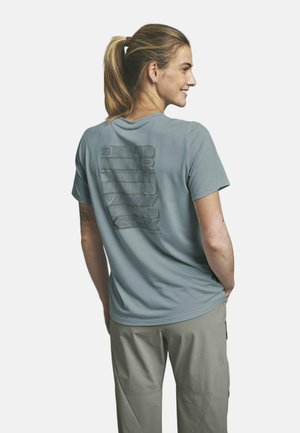 Femme avec une queue de cheval blonde portant un t-shirt graphique bleu clair et un pantalon beige clair, debout les mains dans les poches, souriante, de profil.