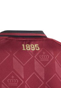 Le maillot de sport rouge présente un col noir avec une fine bande, "1895" imprimé en or, et un motif de couronnes disposées en diamant.