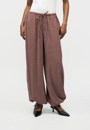 Άτομο που φοράει άνετο, φαρδύ παντελόνι σε αποχρώσεις μαβί με ελαστική μέση και κορδόνι, καθώς και μαύρα τακούνια με μυτερή μύτη, στέκεται μπροστά σε μονόχρωμο φόντο.