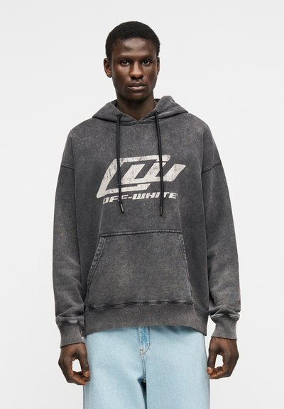 Fiatal férfi, aki egy kifakult fekete Off-White kapucnis pulóvert visel első zsebbel és világoskék farmerral, egyszerű háttér előtt áll.