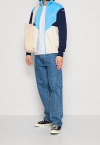 Blå och cremefärgad zip-up jacka med avslappnad passform, kombinerad med ljusblå jeans. Svarta sneakers med vit sula; beige tygväska. Avslappnad sportig stil.