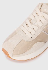 Beige sneaker featuring a mix of suede and mesh materials, flat laces, and a gum rubber sole. Includes stitched accents and a logo.