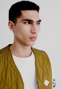 Vest cuquilat de culoare khaki, cu o textură netedă, având o bluză albă cu guler rotund dedesubt. Accesoriu pătrat pe partea stângă a vestului.