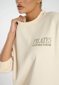 Beige Sweatshirt mit geripptem Ausschnitt und lockerer Passform, mit "PILATES SOUTH BEACH MIAMI" in einer kontrastierenden Farbe bestickt.