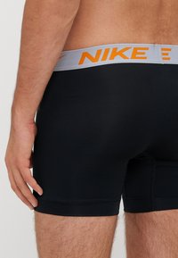 Melni bokserveidā apakšbikses ar pelēku jostasvietu, uz kuras ir oranžs "NIKE" zīmols, ar gludu tekstūru un cieši pieguļošu dizainu.