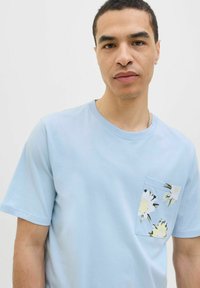 Lyseblå bomulds t-shirt med rund halsudskæring, som har en blomsterprintet lomme på venstre side. Korte ærmer. Glat tekstur.