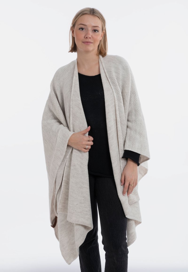 Helle beige Strickponcho mit strukturiertem Muster, offenem Vorderdesign, fallenden Ärmeln und lockerer Passform über einem schwarzen Oberteil und einer Hose.