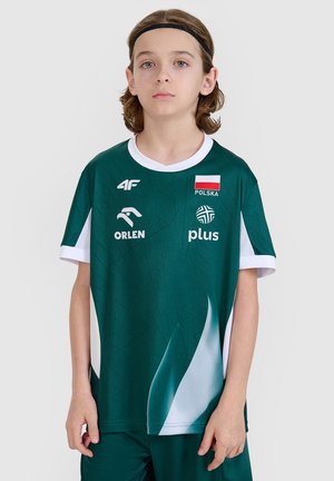 Młoda osoba nosząca zieloną sportową koszulkę Polski z logo, w tym flagą Polski, Orlen, Plus i marką 4F, na jednolitym tle.