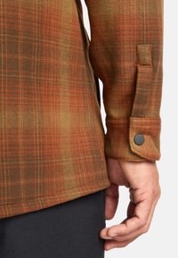 Camisa de franela a cuadros en marrón y naranja, con puños abotonados y tejido texturizado, mostrando un primer plano de la manga y la mano posicionada en la cintura.