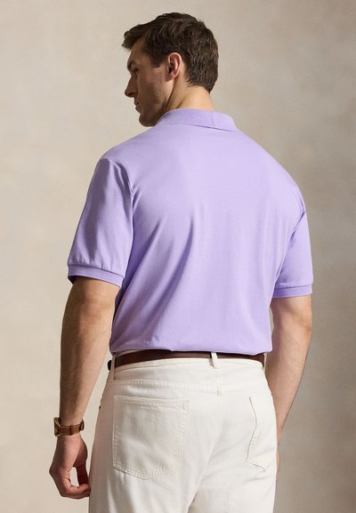 Άνδρας με ανοιχτό μωβ κοντομάνικο πουκάμισο πόλο, κρυμμένο σε λευκό παντελόνι με καφέ δερμάτινη ζώνη, φαίνεται από πίσω.