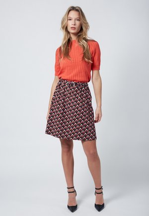Haut rouge à rayures avec des manches courtes associé à une jupe noire à motifs comportant des designs géométriques bleus, roses et blancs. Talons aiguilles noirs.