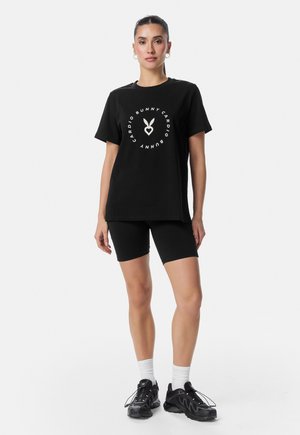 Naine seisab valgel taustal, musta T-särgiga, millel on jänku logo ja tekst, mustad jalgrattashortsid, valged sokid ja mustad tossud.