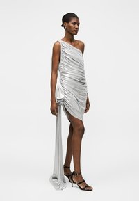 Γυναίκα με ασημί μίνι φόρεμα με έναν ώμο και ραβδώσεις, με μακρύ υφασμάτινο στοιχείο, φορώντας μαύρα ψηλοτάκουνα σανδάλια με λουριά, σε λευκό φόντο.