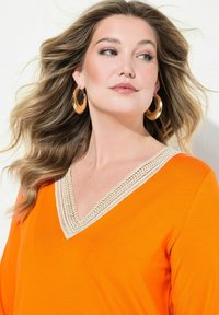 Oranges V-Ausschnitt-Top mit beigem Spitzenbesatz, glatte Textur und langen Ärmeln. Das Model trägt große goldene Creolen, ihr Haar fließt.