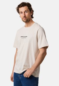 Beige bomulds-T-shirt med en sort trykt logo på brystet. Korte ærmer, rund hals og afslappet pasform. Kombineret med blå jeans.