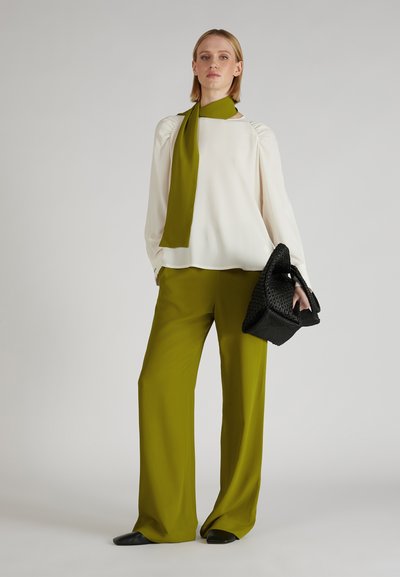 Witte blouse met lange mouwen en opgezette schouders, gecombineerd met olijfgroene wijde broekspijpen. Gekwast met een zwarte gevlochten handtas.