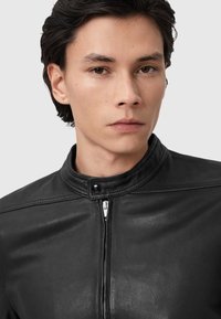 Zwarte leren jas met een hoge kraag, voorzien van een rits aan de voorkant en een drukknoopdetail. Gladde textuur en getailleerde pasvorm.