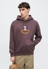 Sweat à capuche violet avec une poche avant et un cordon à la capuche, orné d'un graphisme représentant un personnage et le logo "Volcom" aux couleurs vives.