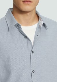 Chemise texturée bleu clair avec un col boutonné, présentant des boutons foncés et des détails de col intérieur blanc.