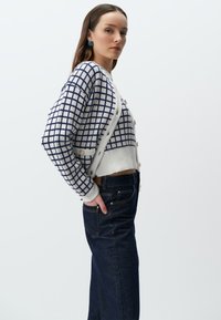 Gebreide cardigan in een wit en marineblauw ruitpatroon, met een knoopsluiting aan de voorkant en een geribbelde zoom. Gecombineerd met donkere denim jeans, met handen in de zakken.