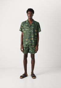 Grøn kortærmet skjorte- og shorts-sæt lavet af letvægtsstof, med et mønsterdesign og forskellige illustrationer. Sorte sandaler.