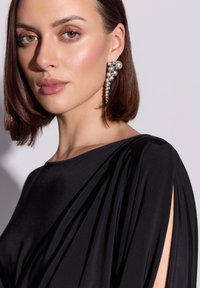 Czarna sukienka z drapowaniem, wycięciami na ramionach i gładką fakturą. Modelka nosi efektowną srebrną kolczyk z okrągłymi koralikami zwisającymi w dół.