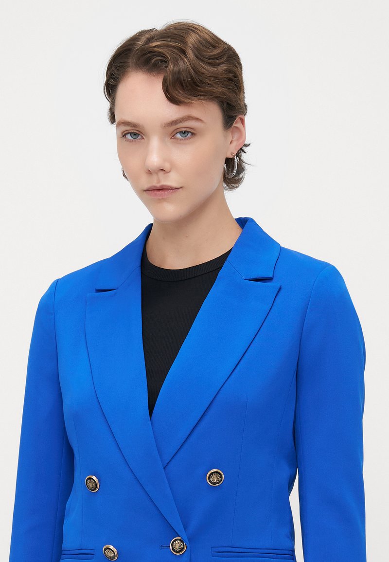 Blazer ajusté bleu vif avec un design à double boutonnage, des revers crantés et des accents en boutons dorés. Porté sur un haut noir. Texture lisse.