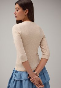 Beżowa, prążkowana bluzka z długim rękawem o dopasowanym kroju, zestawiona z wielowarstwową spódnicą dżinsową. Widok od tyłu ukazuje rękawy do łokcia oraz subtelną fakturę.