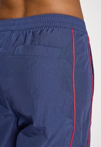 Närbild av en person som bär navyblå sportshorts med röda sidosträck och elastisk midja mot en vit bakgrund.